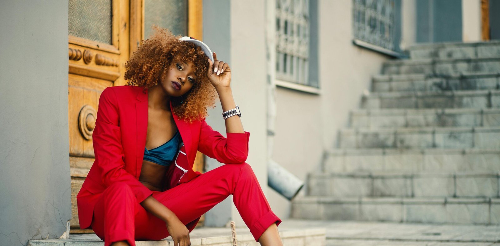 A fashionable young woman in a vibrant red suit poses outdoors on stairs in an urban setting.